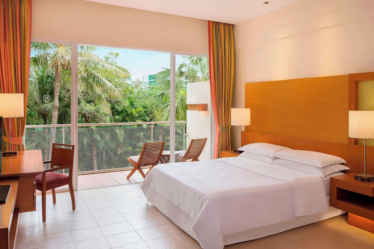 Une chambre d’hôtel avec un grand lit, un bureau et des chaises. Elle dispose d’un balcon avec vue sur les palmiers et les immeubles.