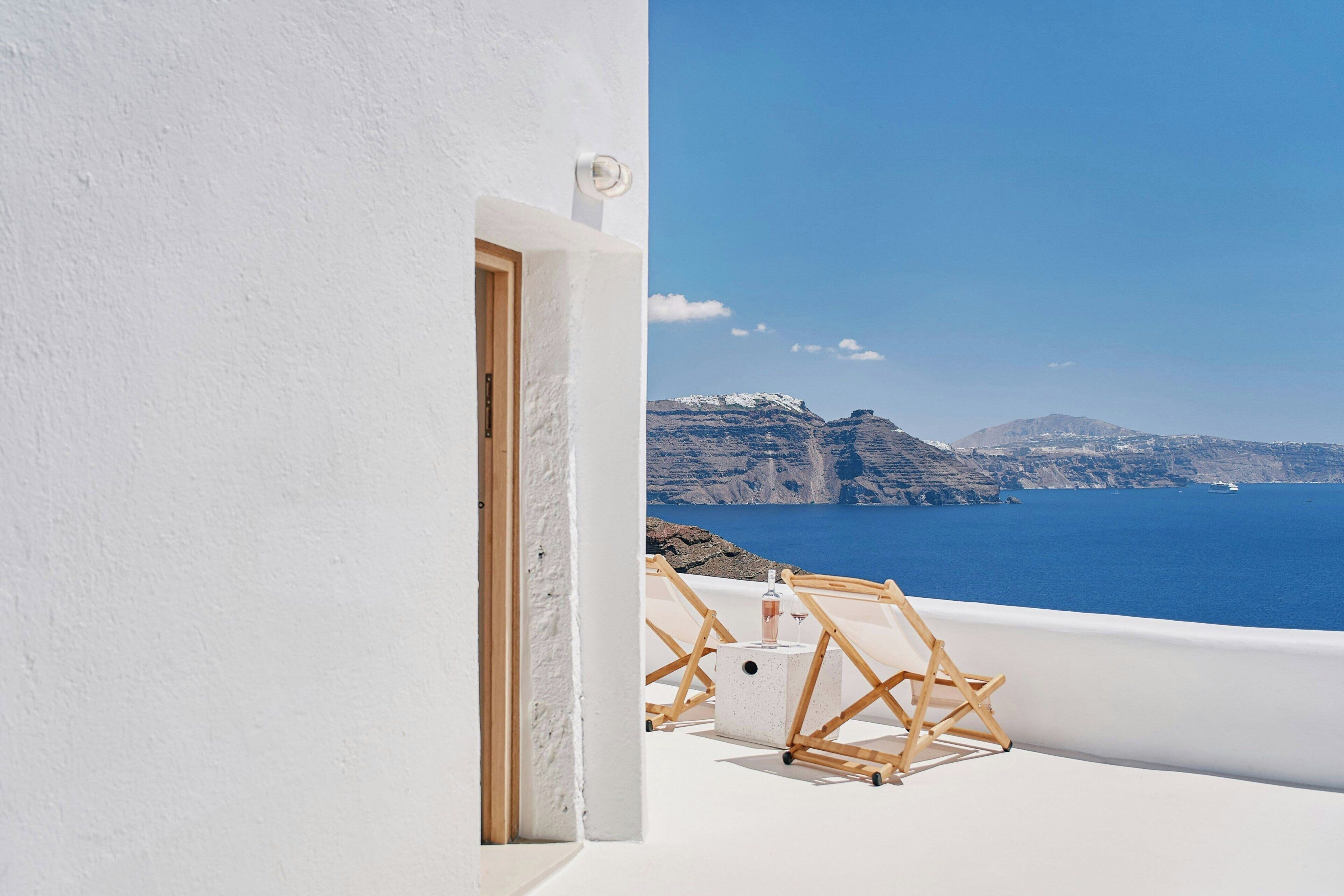 A white building with a door leading to a balcony overlooking a blue sea and distant mountains.