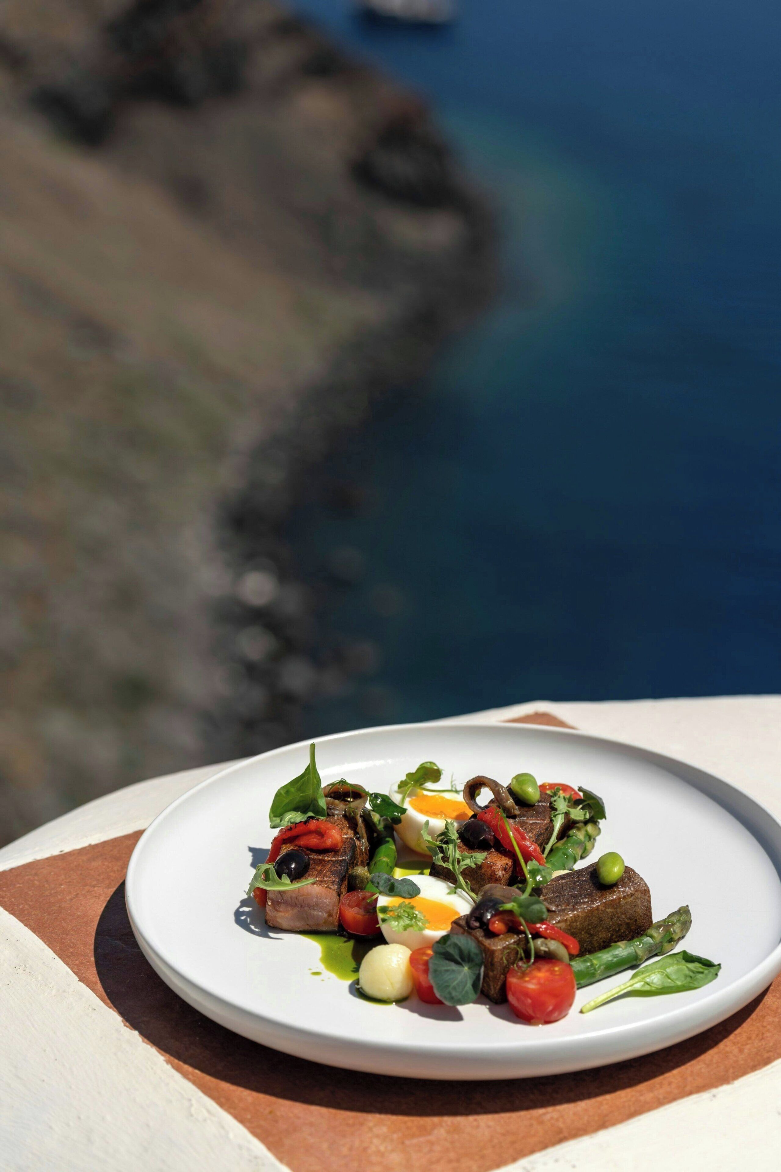 Une assiette blanche, sur laquelle sont disposés divers aliments, est posée sur une terrasse offrant une vue sur la mer.