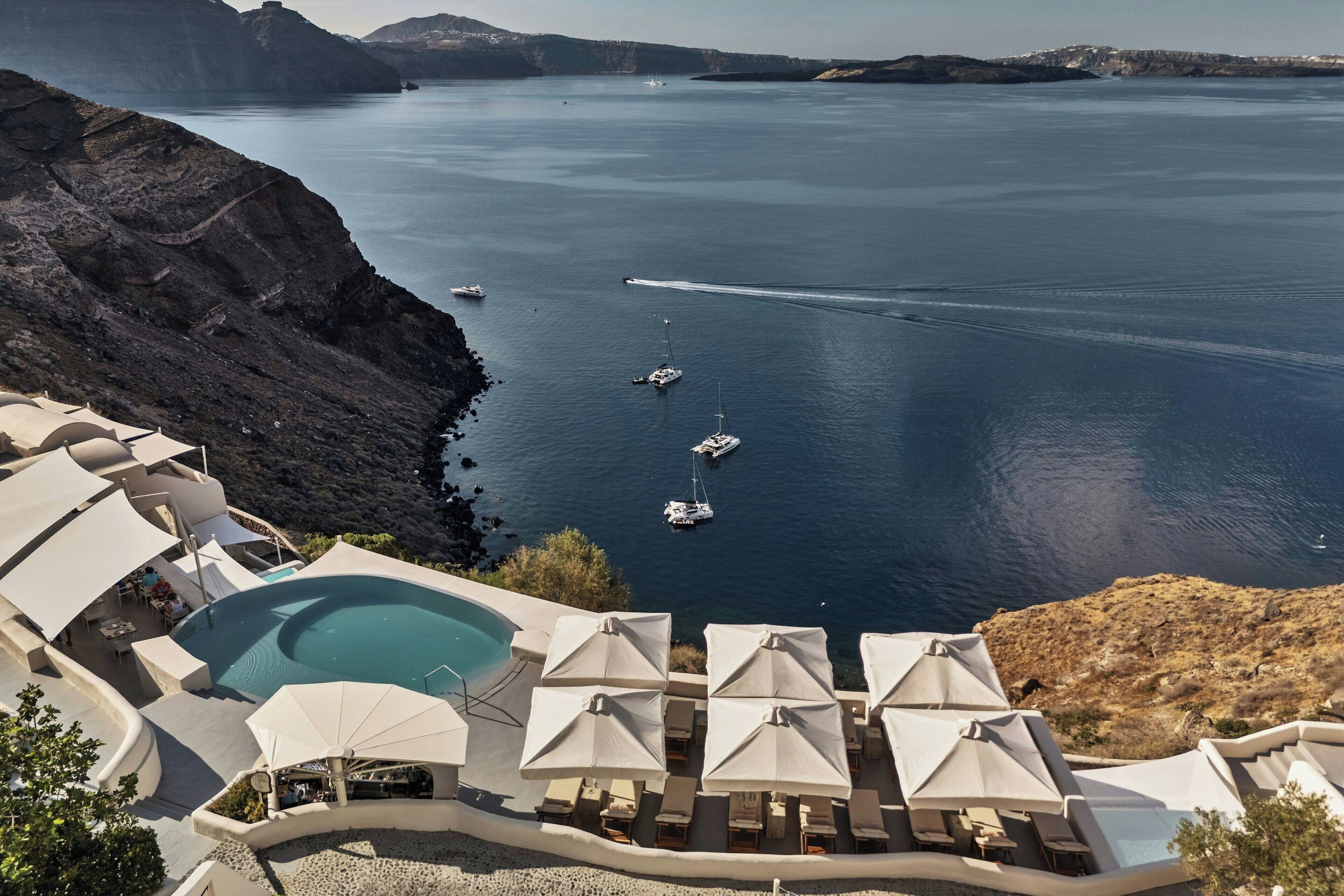 A resort with a swimming pool, lounge chairs, and umbrellas, overlooking a calm sea with boats.