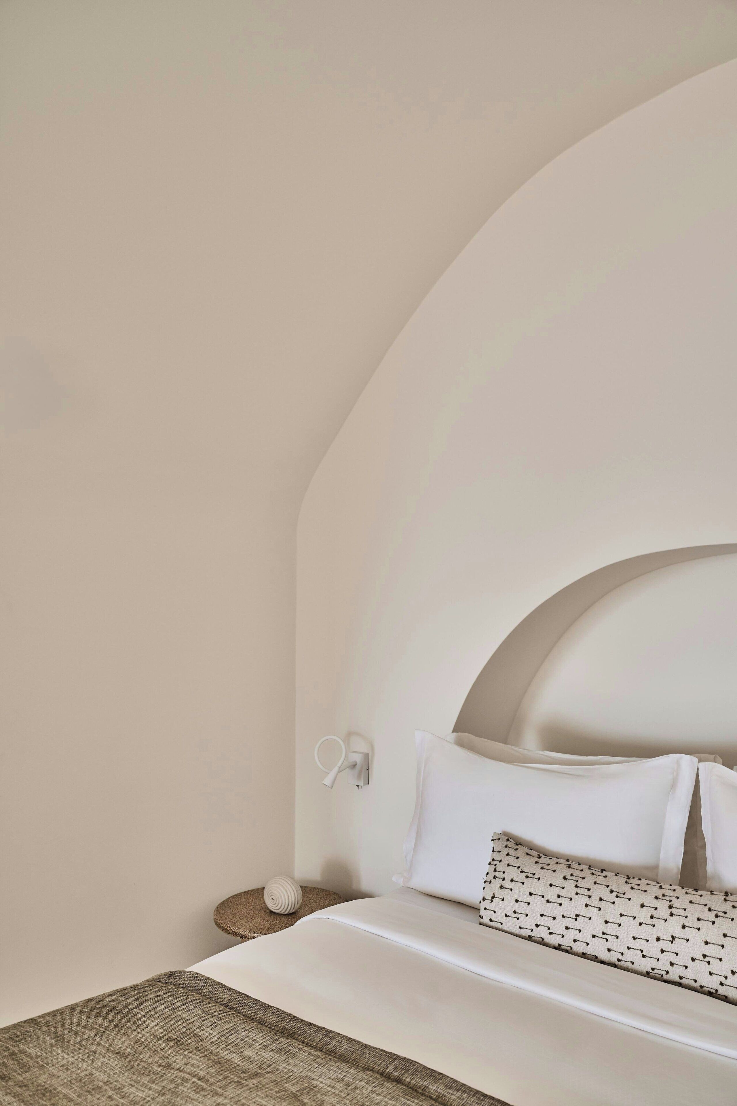 A neatly arranged bedroom with a curved ceiling, a bed with white linens, a patterned pillow, and a small round table with a white object.