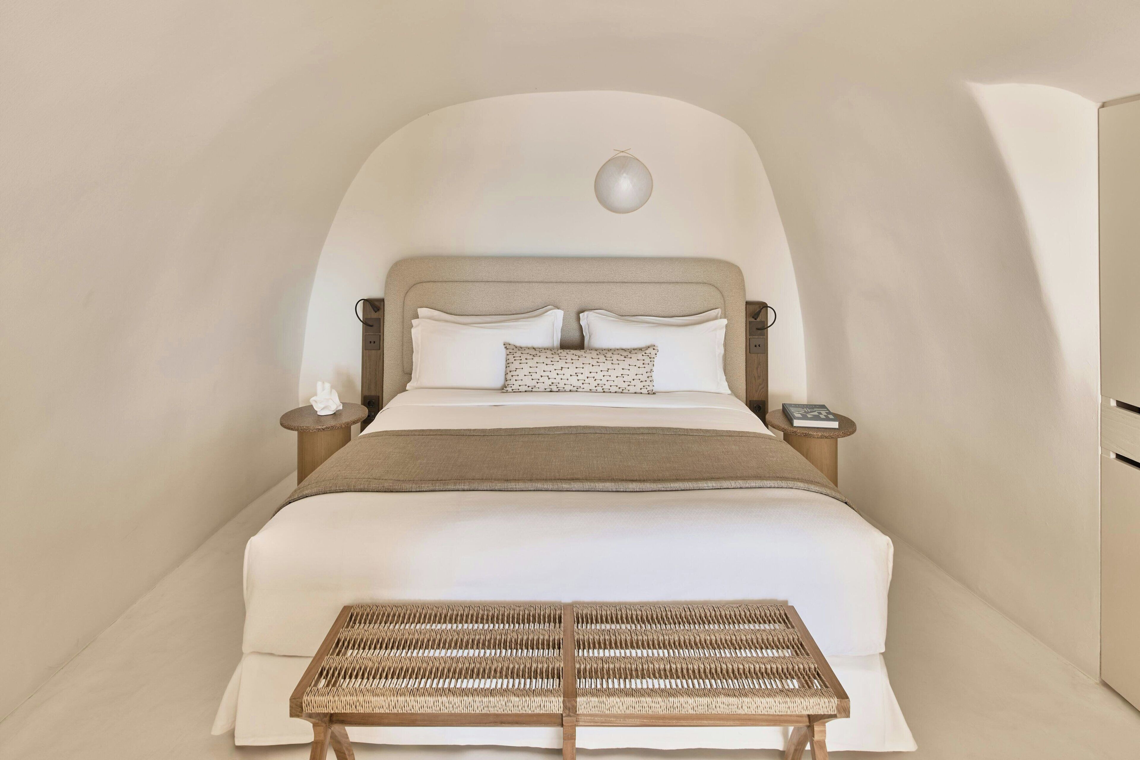 A spacious bedroom with a large bed, a bedside table, a small round table, and a woven bench.