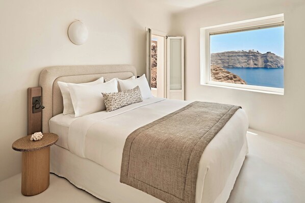 A bedroom with a bed, a bedside table, a nightstand, a window with a view of cliffs and water, and a door leading to another room.