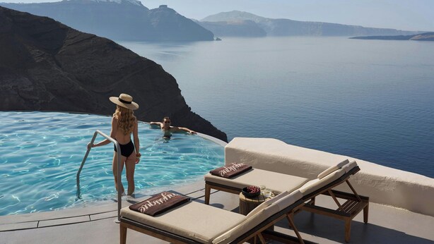 An infinity pool with a view of the sea and mountains, lounge chairs with towels, and a person in the water. - Mystique, a Luxury Collection Hotel, Santorini (Santorini)
