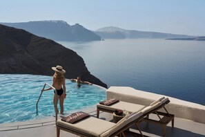 An infinity pool with a view of the sea and mountains, lounge chairs with towels, and a person in the water.