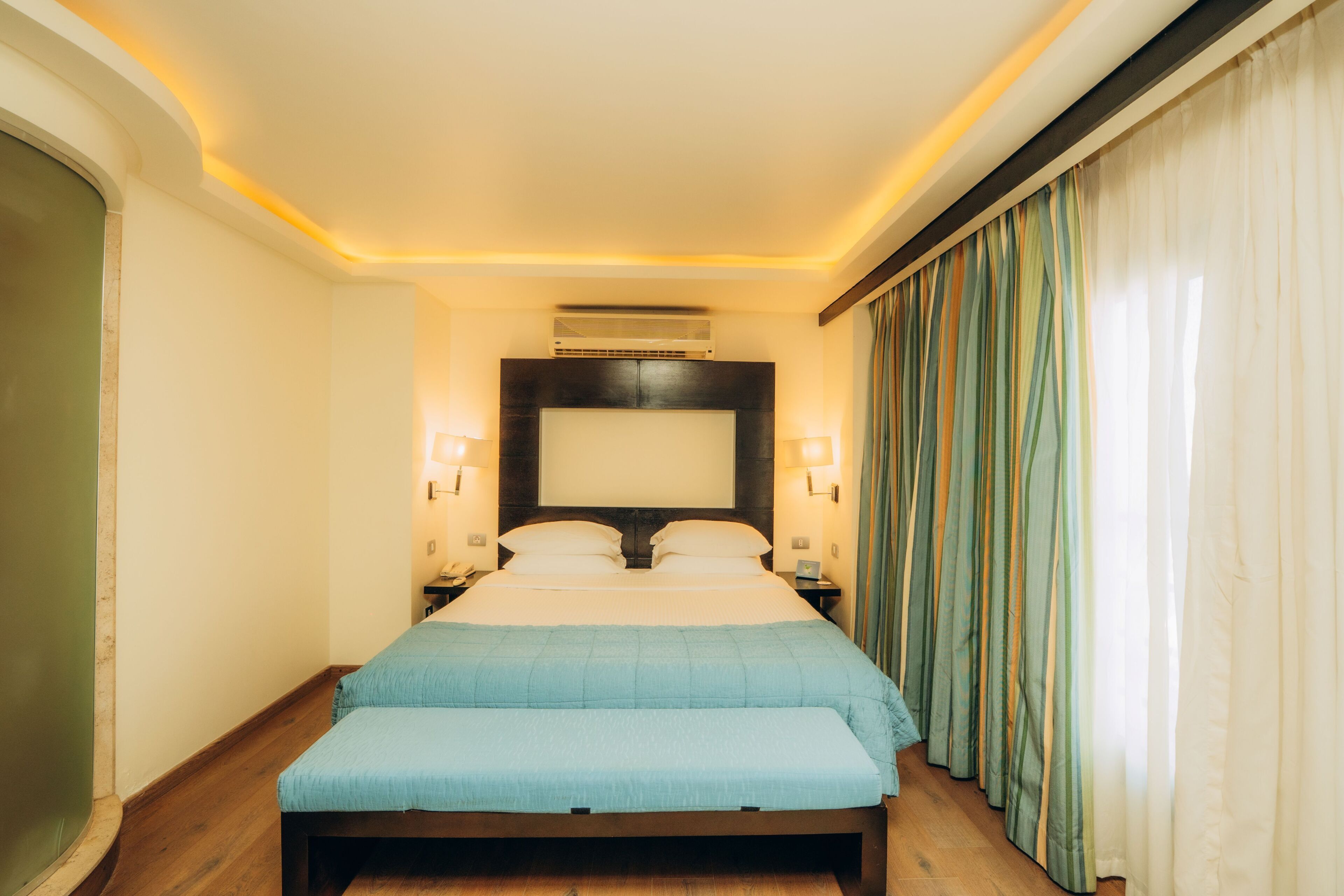 A hotel room with a bed, bedside lamps, an air conditioning unit, a nightstand, and a curtain.