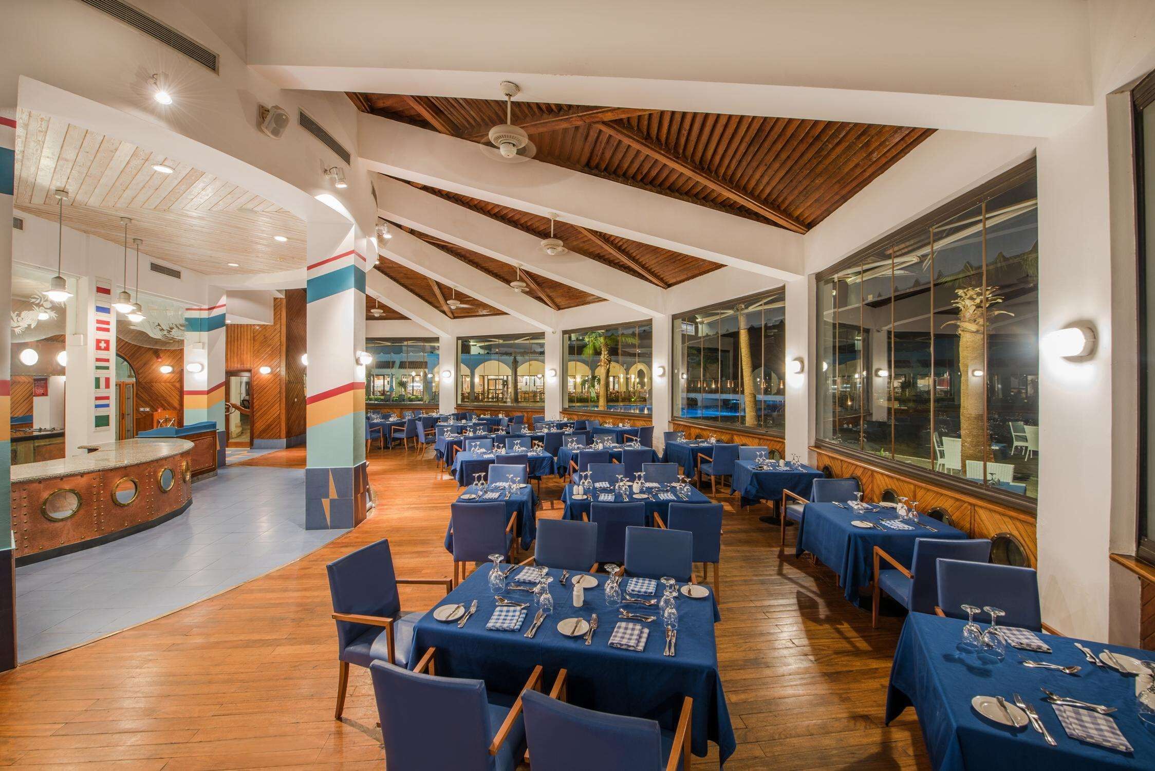 A spacious dining area with multiple tables set for guests, featuring a high ceiling with wooden beams and large windows offering views of the outdoors.
