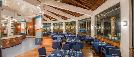 A spacious dining area with multiple tables set for guests, featuring a high ceiling with wooden beams and large windows offering views of the outdoors.