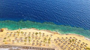 Eine Vogelperspektive zeigt einen Strand mit Liegestuhlreihen und Palmen, ein klares blaues Meer und einen Sandstrand.