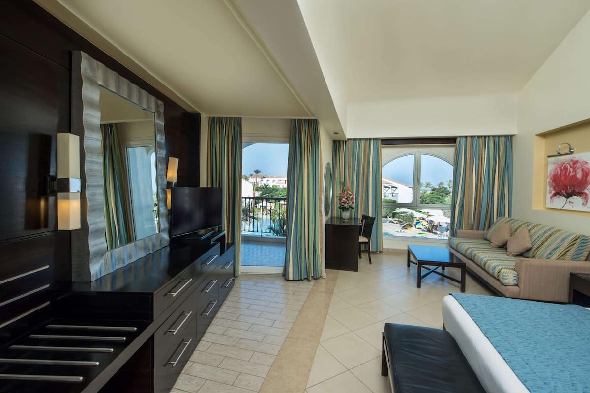 A modern hotel room with a large bed, a TV, a dining area, and a balcony with curtains.