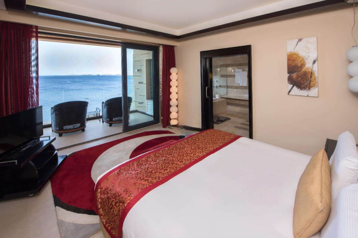 A hotel room with a bed, a TV, a balcony with a view of the sea, and a painting on the wall.
