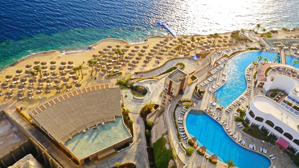 Eine Luftaufnahme eines Resorts mit einem großen Pool, zahlreichen Liegestühlen und einem Strandbereich mit Sonnenschirmen.