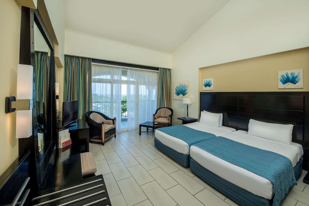 A hotel room with two beds, a desk, a chair, a TV, and a view of the outdoors.