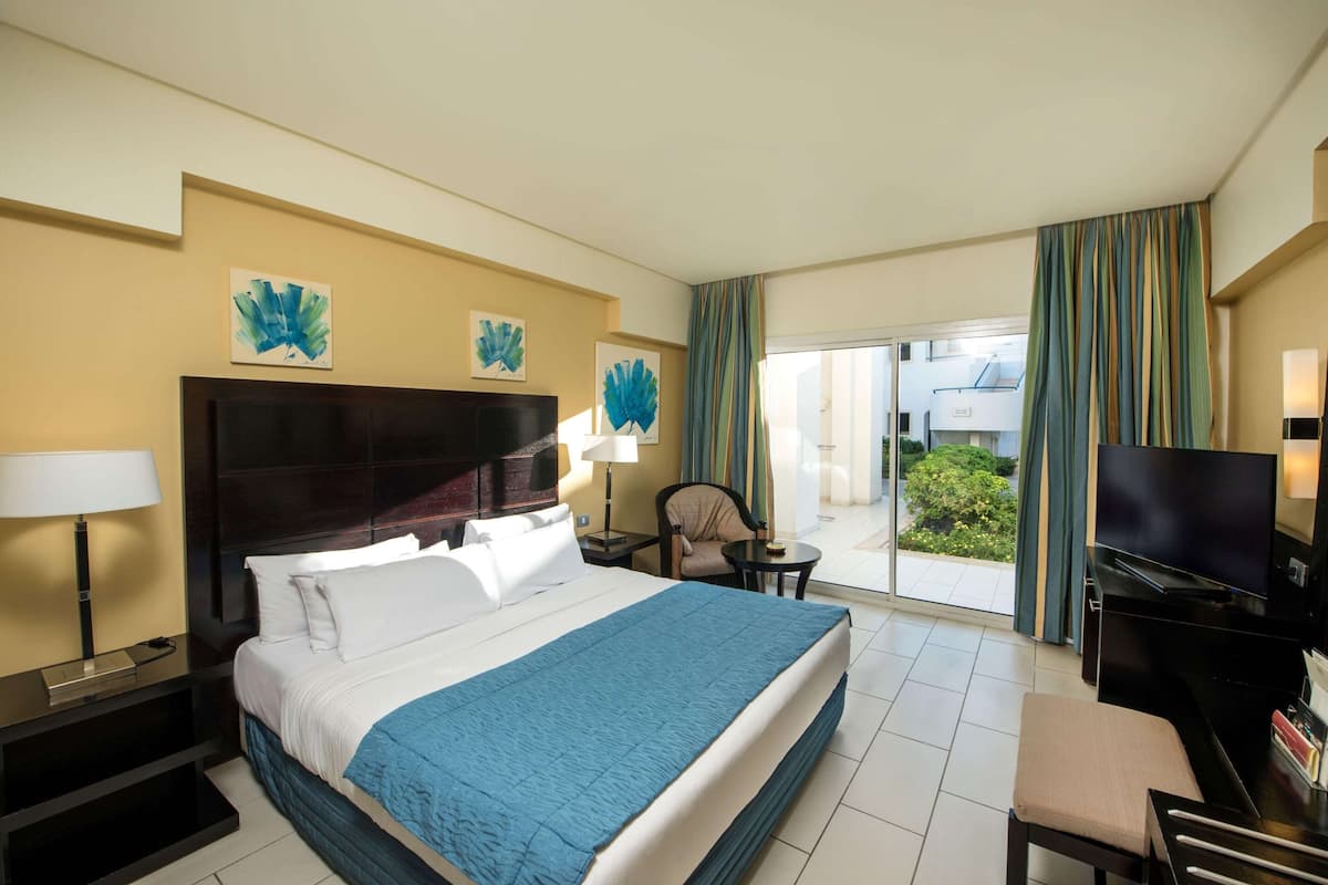 A hotel room with a large bed, a TV, a desk with a chair, and a balcony with curtains.