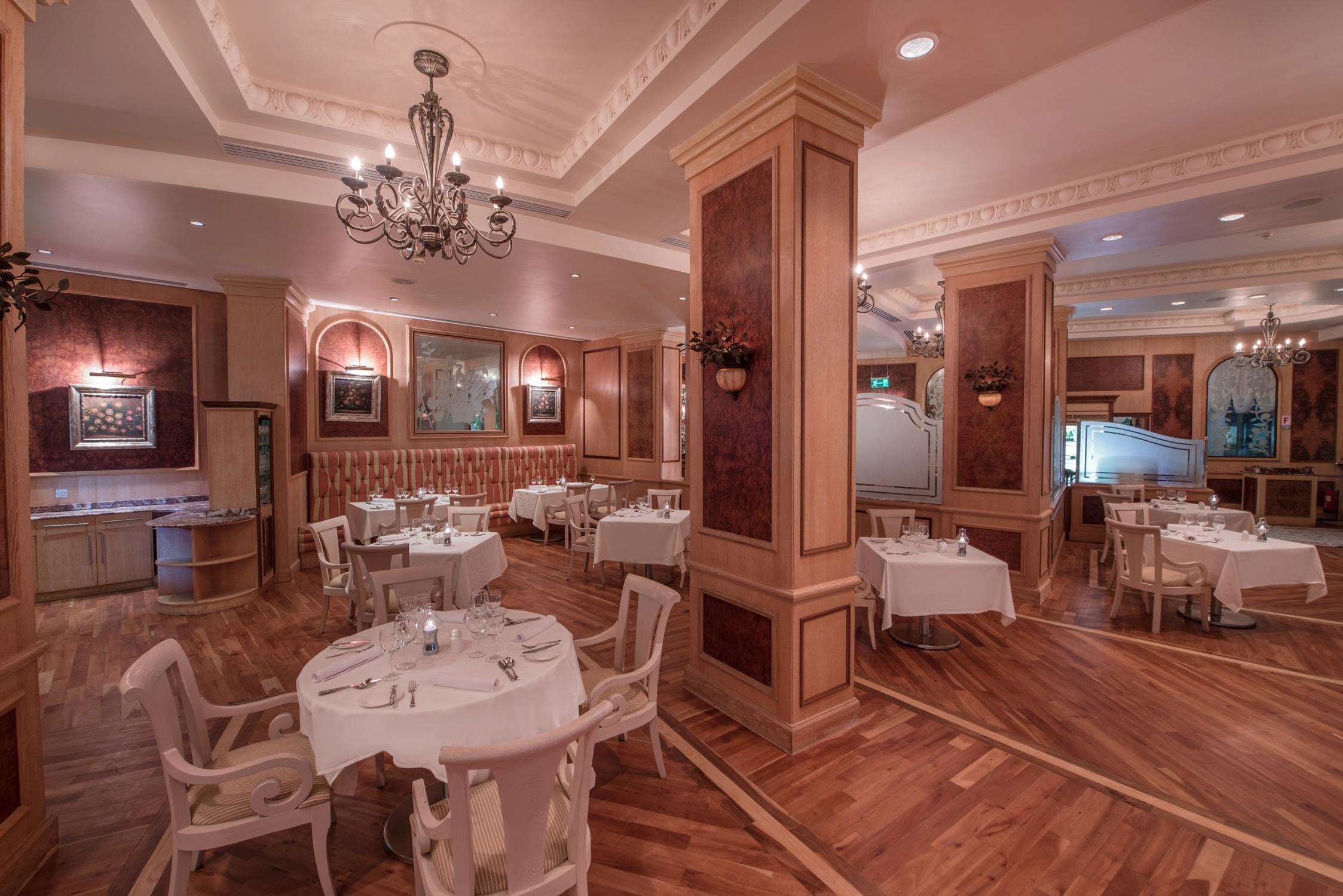 A spacious dining area with multiple round tables and white chairs, wooden paneling, and a chandelier.