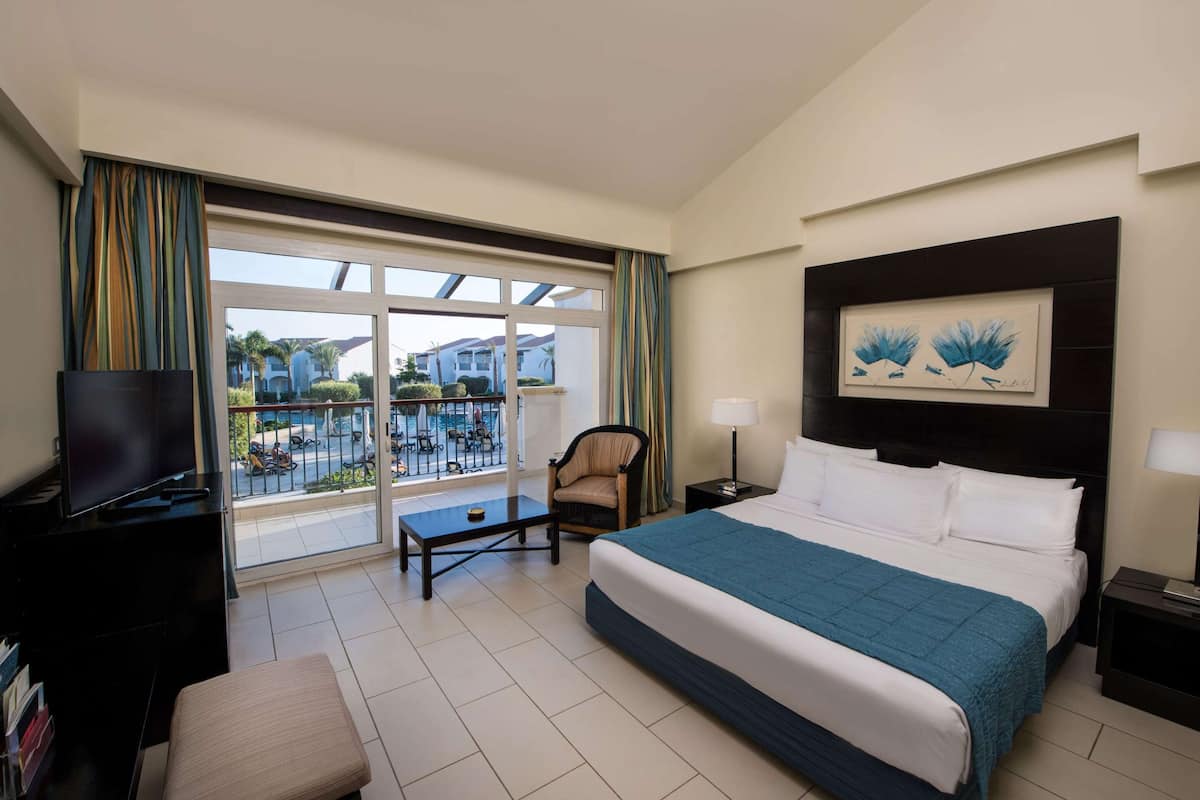 A hotel room with a bed, a TV, a chair, a desk, and a balcony with a view of the pool and buildings.