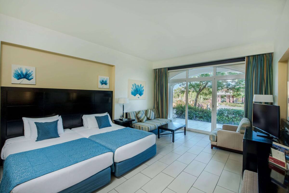 A hotel room with two beds, a sofa, a TV, and a balcony with a view of greenery.