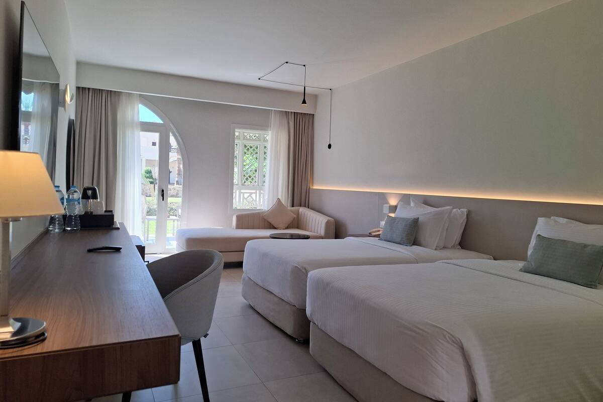 A hotel room with a desk, two beds, a chair, a lamp, and a view of the outdoors.