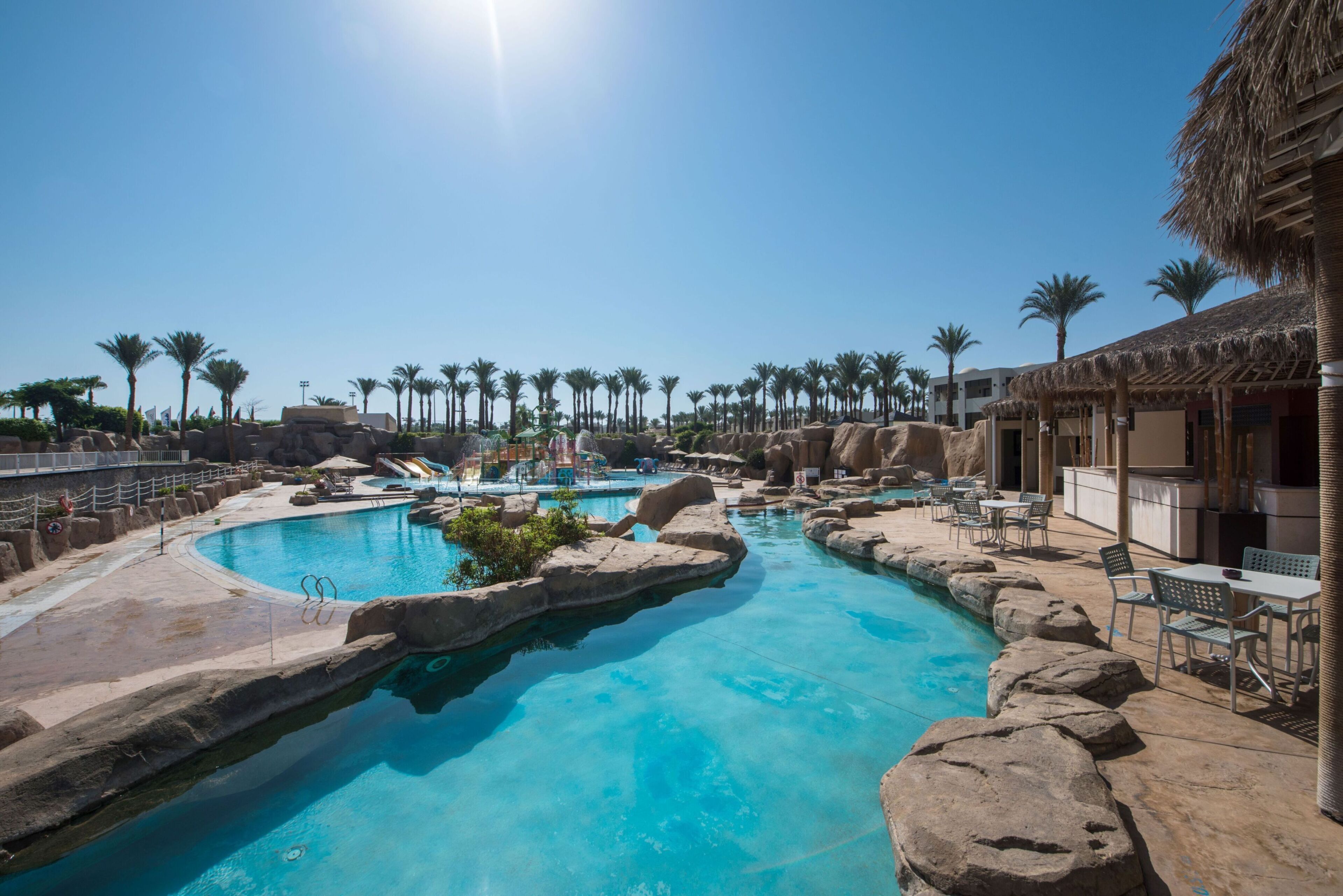 Ein Resort-Pool mit einem klaren, blauen Wasserspiel, umgeben von Palmen und einem Gebäude mit einem Reetdach.