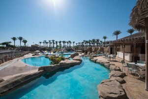 A resort pool with a clear blue water feature, surrounded by palm trees and a thatched-roof building.
