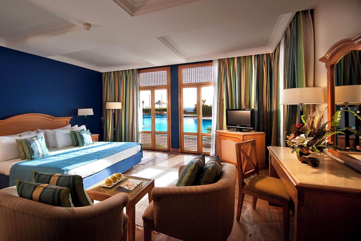 A hotel room with a large bed, a desk with a TV, a chair, and a view of a swimming pool.