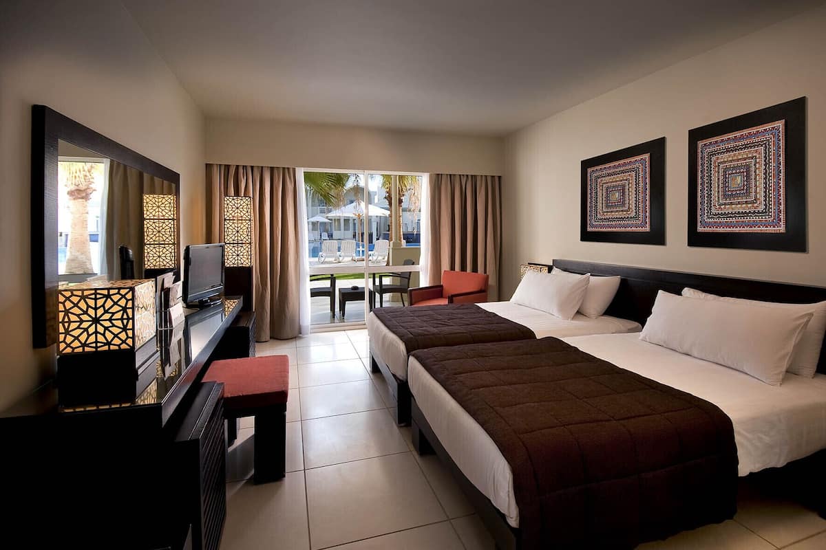 A hotel room with two beds, a desk with a TV, and a view of the outside.