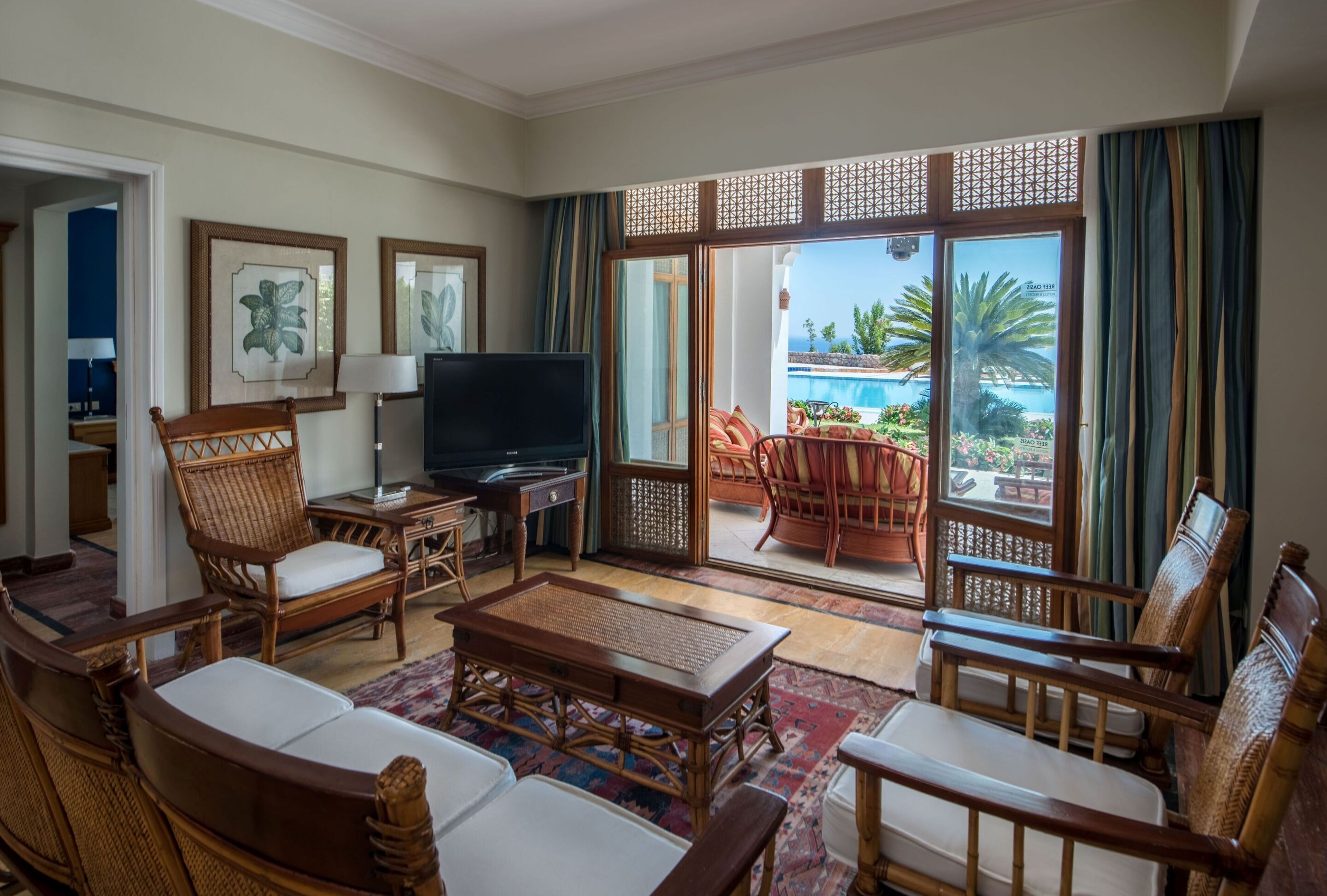 Amplia sala de estar con mobiliario de mimbre, televisor de pantalla plana y vista a una piscina y palmeras.