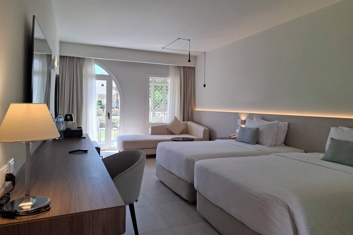 A hotel room with a desk, two beds, a chair, a mirror, and a window with curtains.
