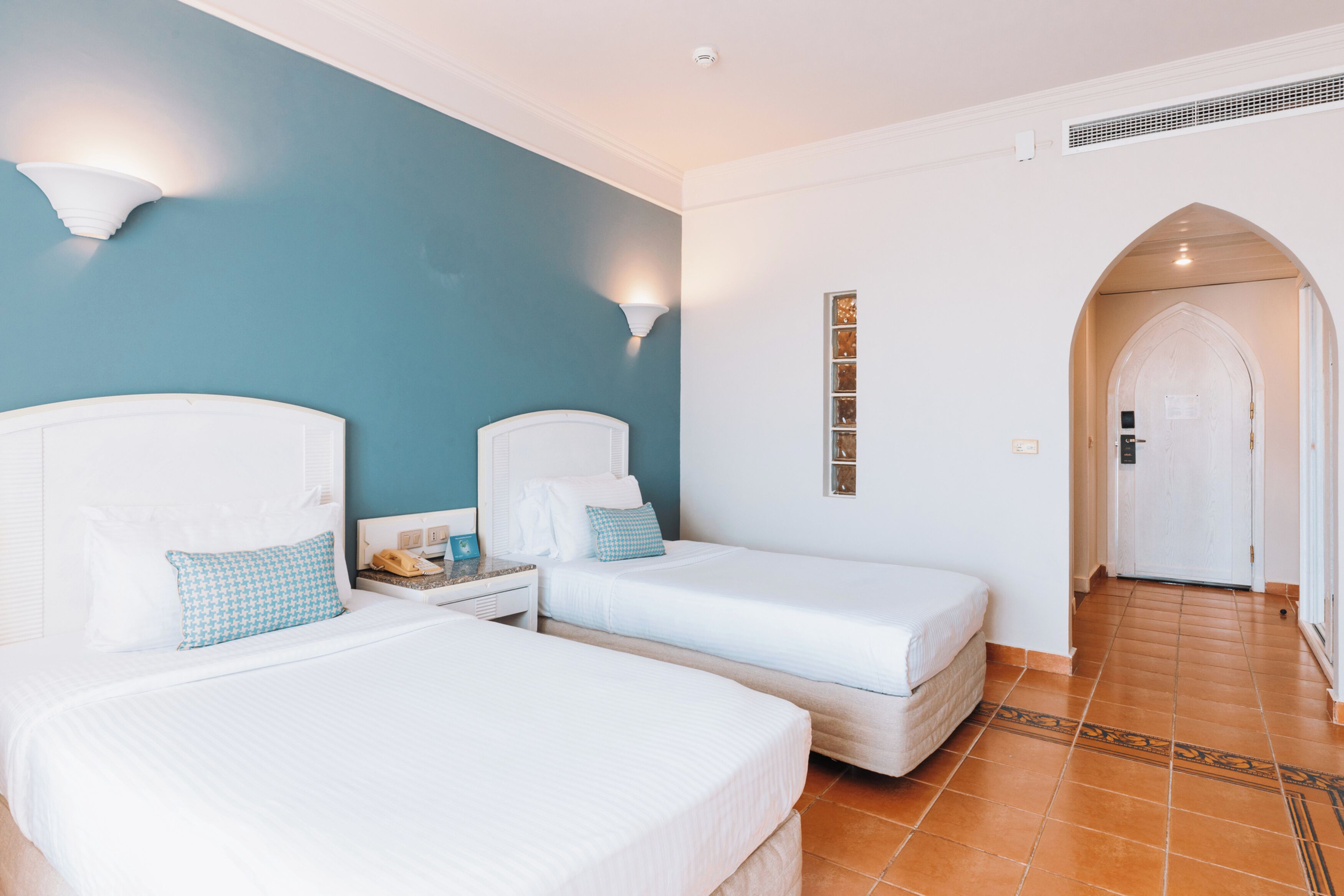 A hotel room with two beds, a nightstand, a wall-mounted lamp, and a door leading to another room.