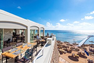 A seaside restaurant with outdoor seating overlooking the ocean, featuring tables and chairs, and a view of the beach.