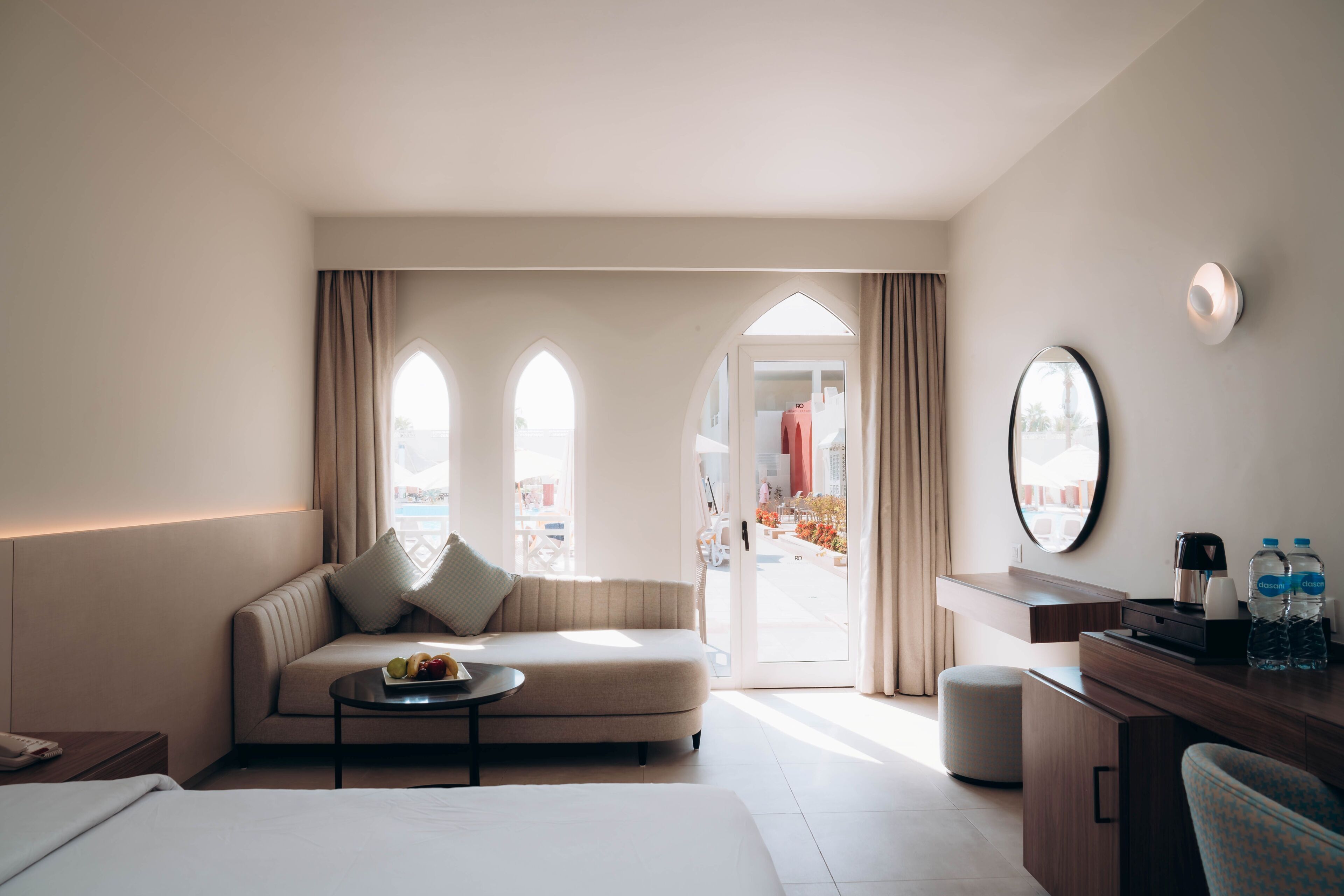 Habitación de hotel con cama, sofá, mesa de centro con fruta, escritorio con espejo y vistas al exterior.