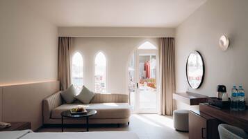 Habitación de hotel con cama, sofá, mesa de centro con fruta, escritorio con espejo y vistas al exterior.