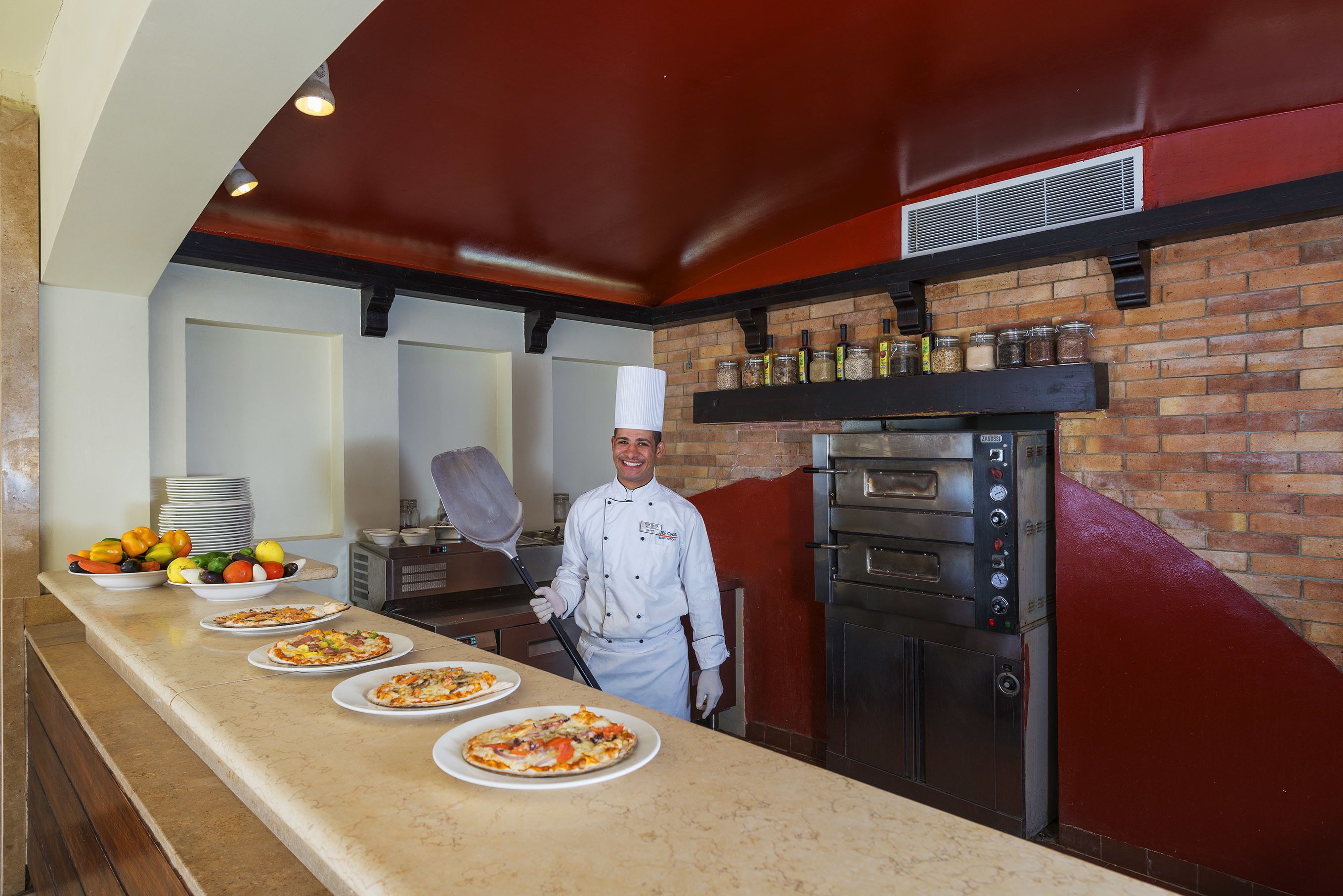 Un chef de cocina en una cocina con pared de ladrillo, horno de pizza y mostrador con pizzas.
