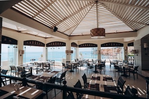 A spacious dining area with multiple tables and chairs, large windows offering a view of the sea, and a high ceiling with a patterned ceiling fan.