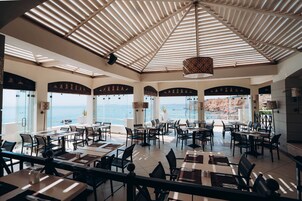 A spacious dining area with multiple tables and chairs, large windows offering a view of the sea, and a high ceiling with a patterned ceiling fan.