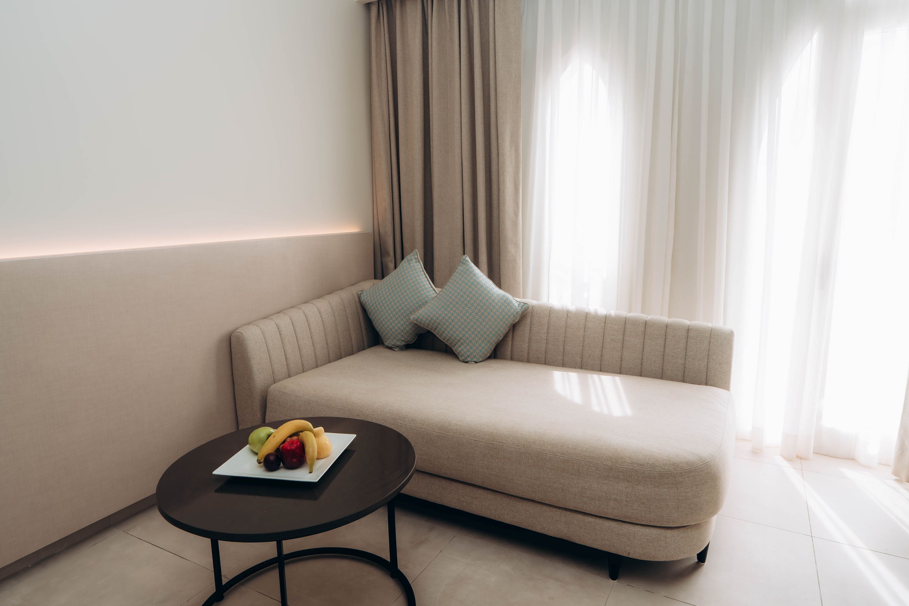 Una sala de estar moderna con un sofá beige, dos almohadas estampadas, una mesita redonda con una bandeja de frutas y cortinas de gasa.