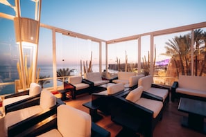 Un salón moderno con sofás blancos y mesas, grandes ventanales con vistas al mar y al paisaje urbano.