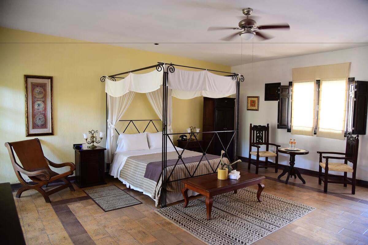 A four-poster bed with a canopy, a ceiling fan, a wooden chair, a small table with a vase, and a framed picture on the wall.