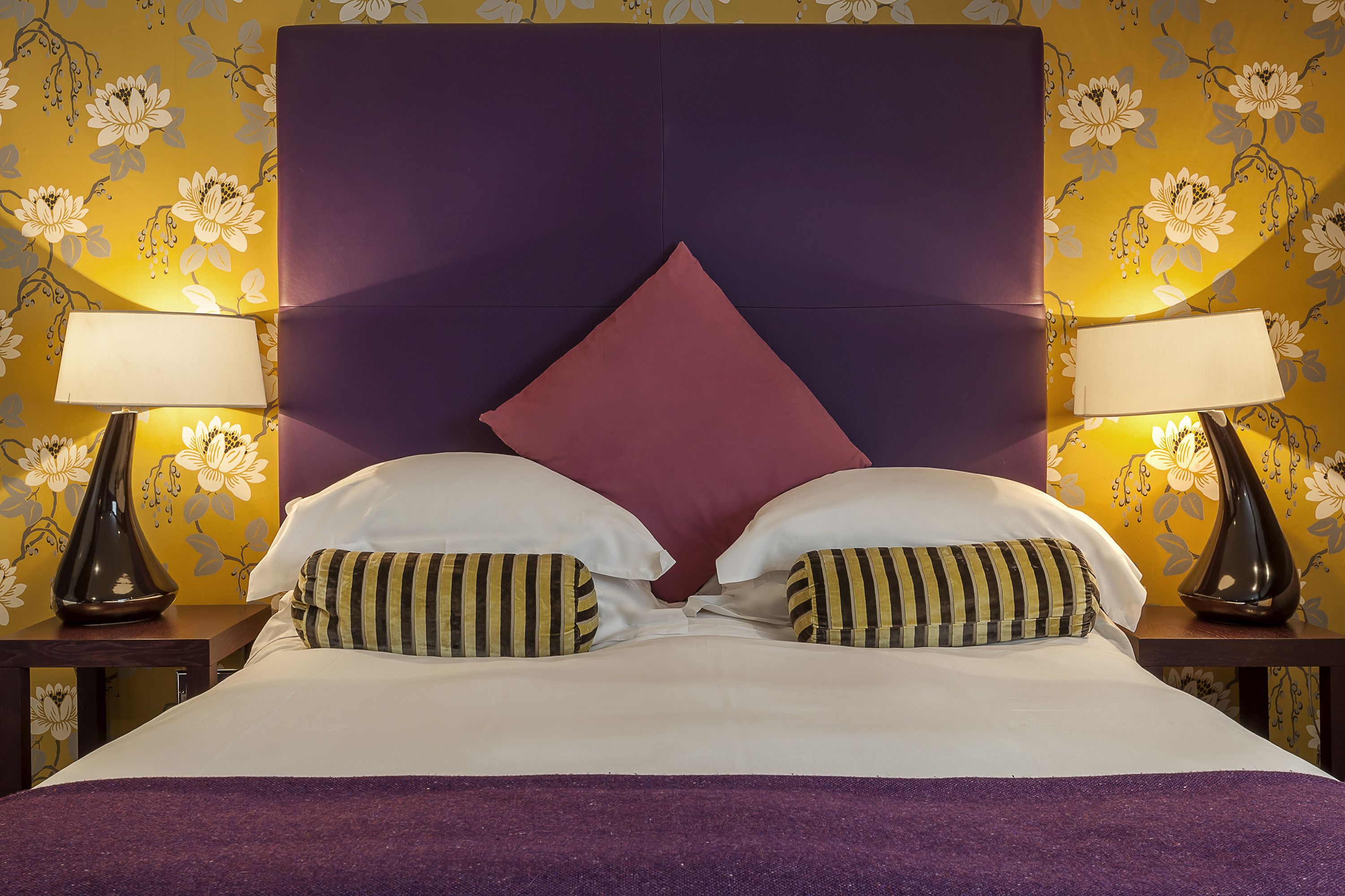 Una cama con cabecera morada, dos almohadas blancas y almohadas a rayas. Hay dos lámparas a ambos lados de la cama.