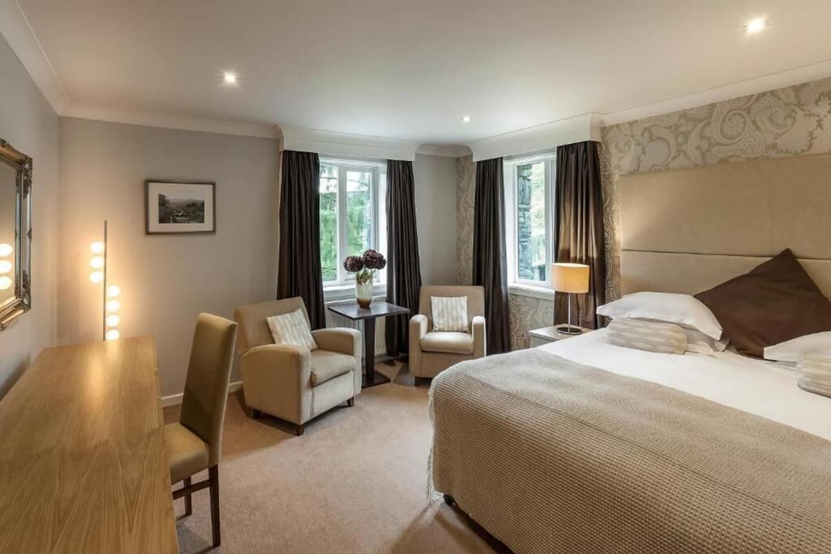 A hotel room with a large bed, two armchairs, a small table with a vase, and a desk with lights.