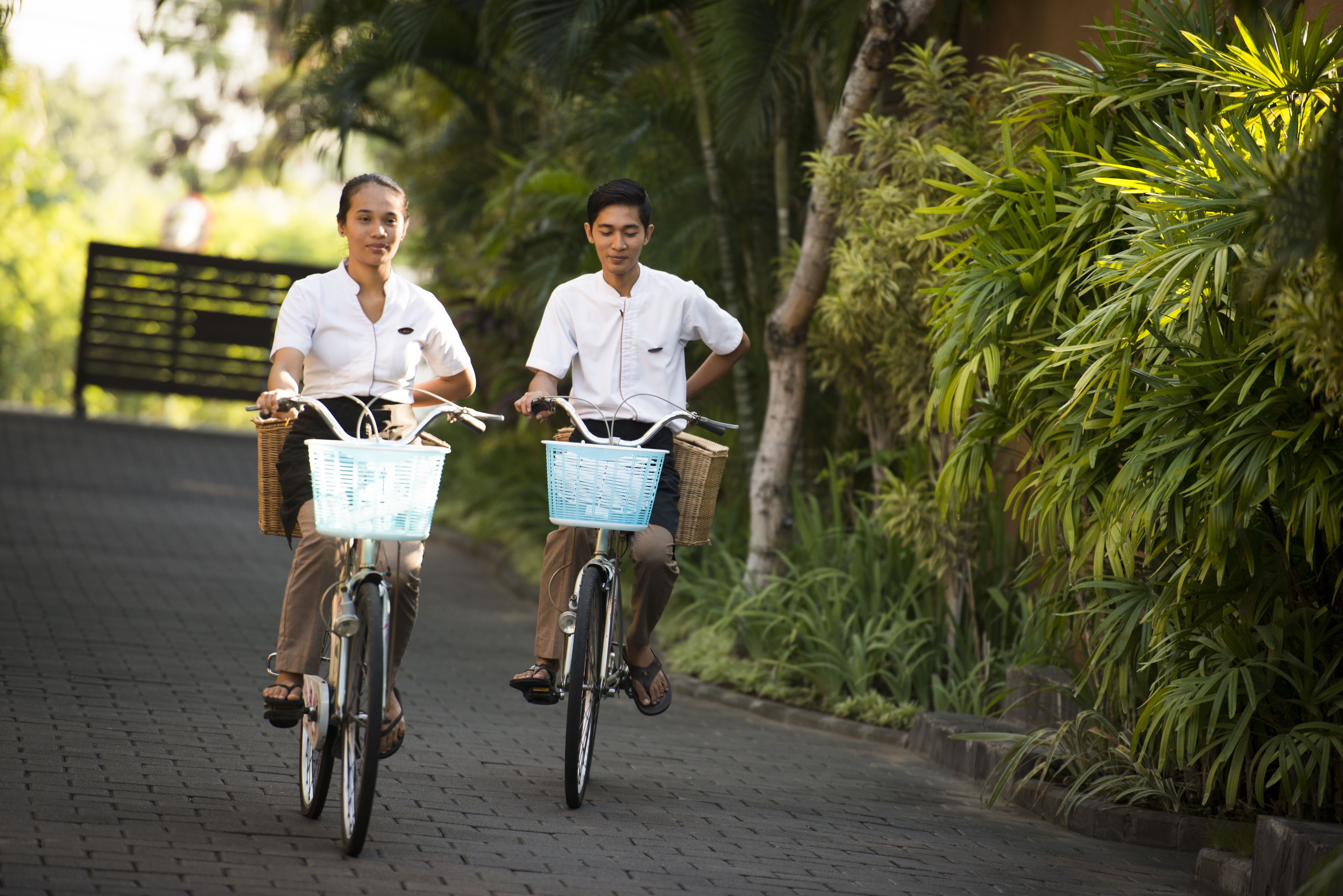 Two people riding bicycles on a paved path with greenery on the sides.