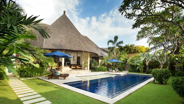 A pool area with a thatched-roof building, lounge chairs, and umbrellas. - The Kunja (Seminyak)