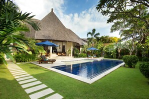 A pool area with a thatched-roof building, lounge chairs, and umbrellas. - The Kunja (Seminyak)