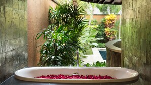 A bathroom with a bathtub filled with rose petals, surrounded by lush green plants and a stone wall. - The Kunja (Seminyak)