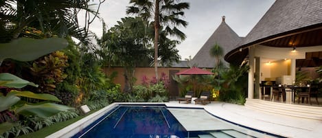 Ein Swimmingpool, umgeben von üppiger tropischer Vegetation, einer strohgedeckten Struktur und einem Essbereich mit Sitzgelegenheiten im Freien.