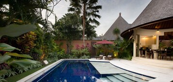 Une piscine entourée d’une végétation tropicale luxuriante, une structure à toit de chaume et un espace repas avec des sièges en plein air.