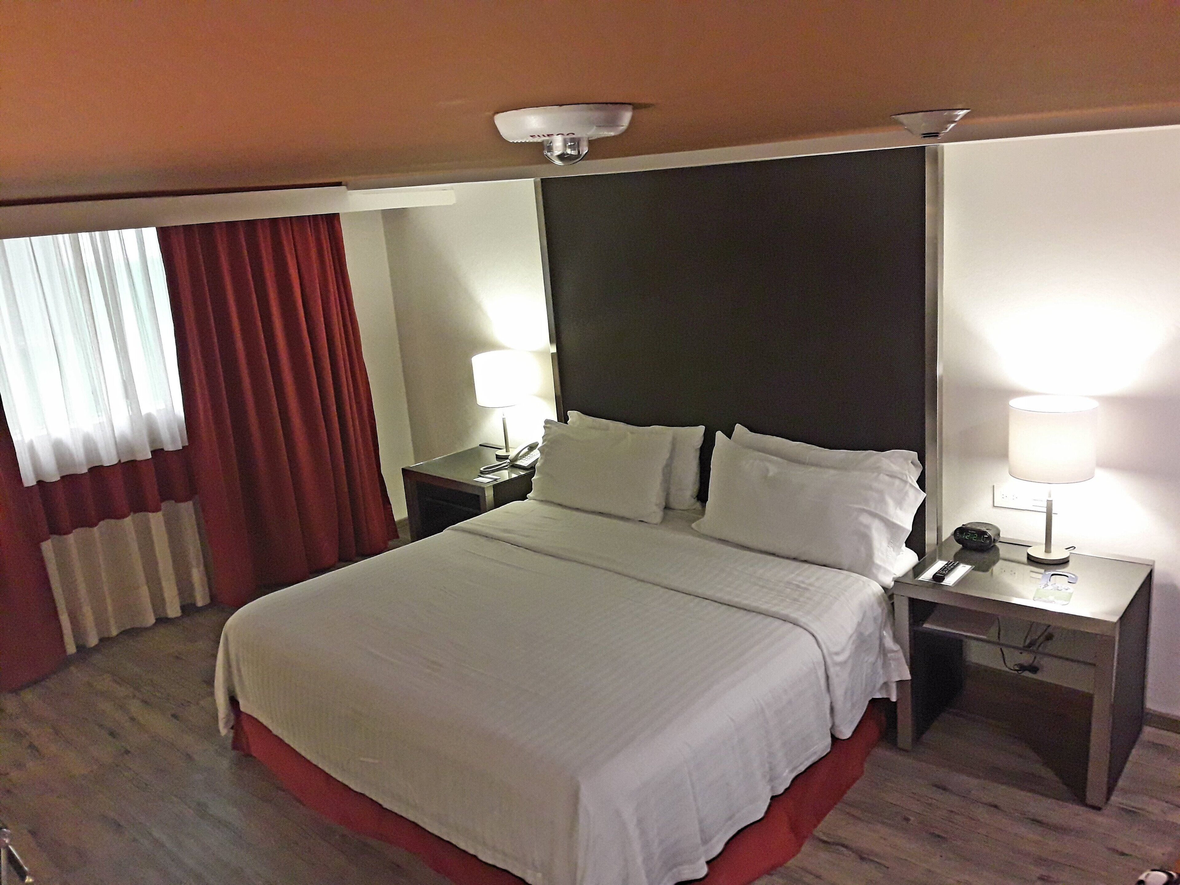 Habitación de hotel con cama, dos mesitas de noche, lámpara y espejo.
