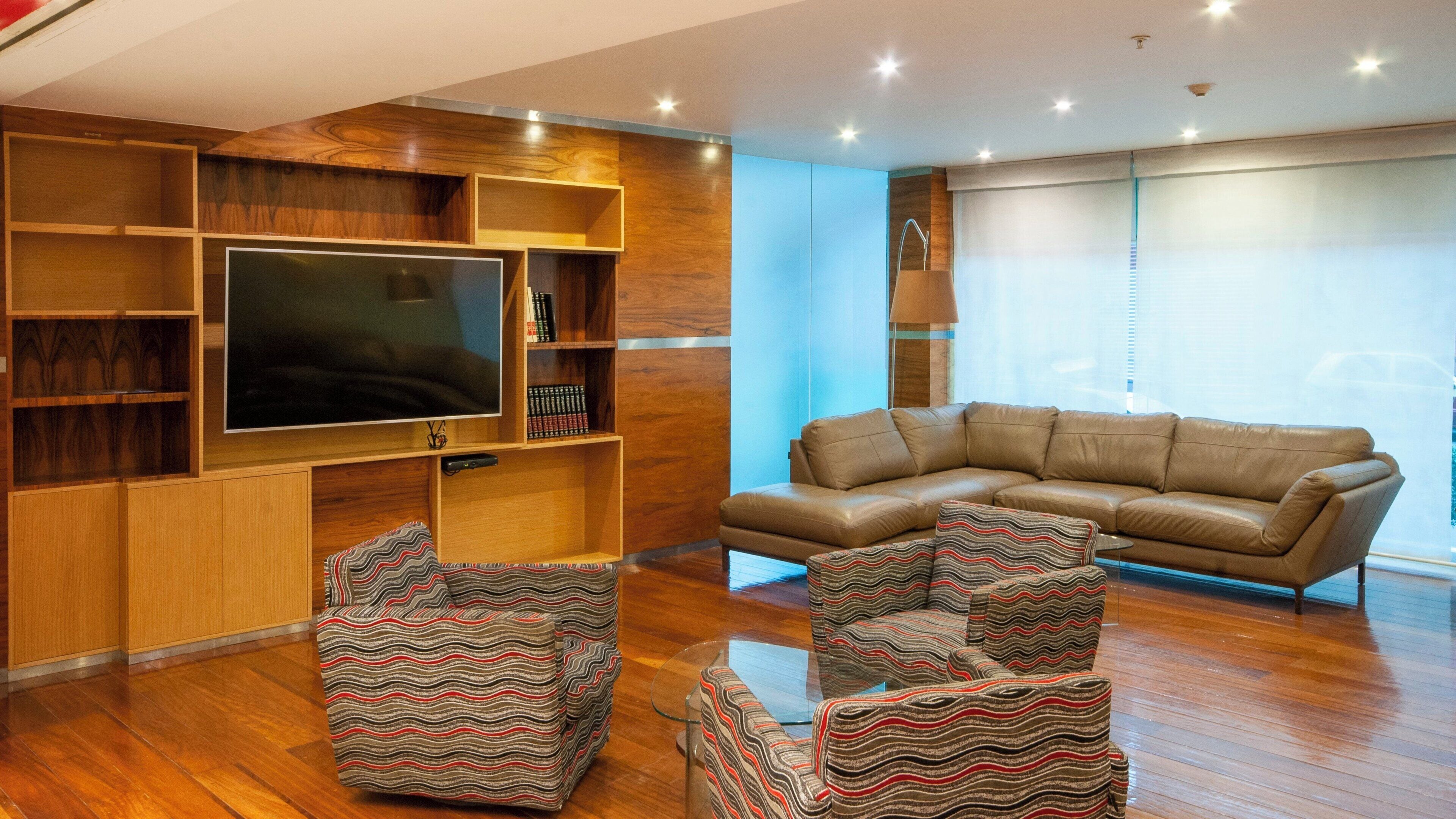 A modern living room with a large flat-screen TV, wooden shelving, a leather sofa, and patterned armchairs.
