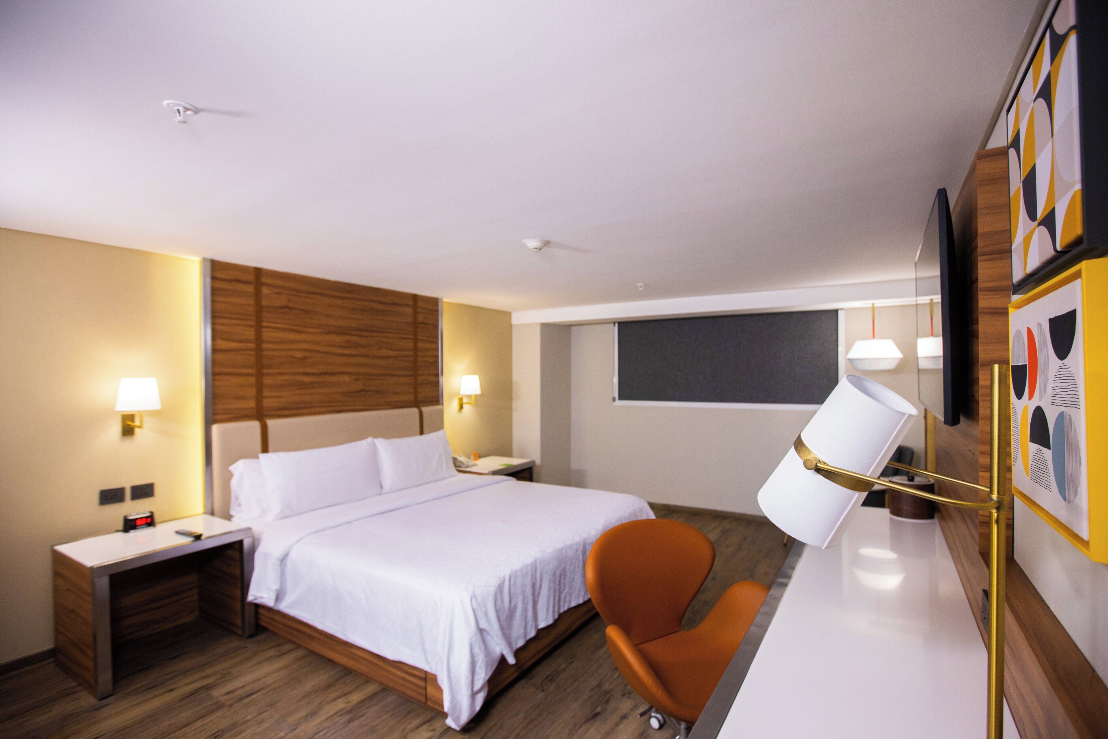 Habitació d'hotel amb un llit, un escriptori, una cadira taronja i una làmpada penjada a la paret.