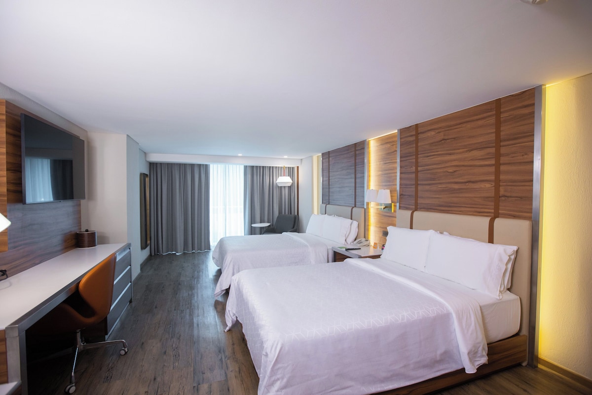 Een hotelkamer met twee bedden, een bureau, een stoel, een lamp en een groot raam met gordijnen.